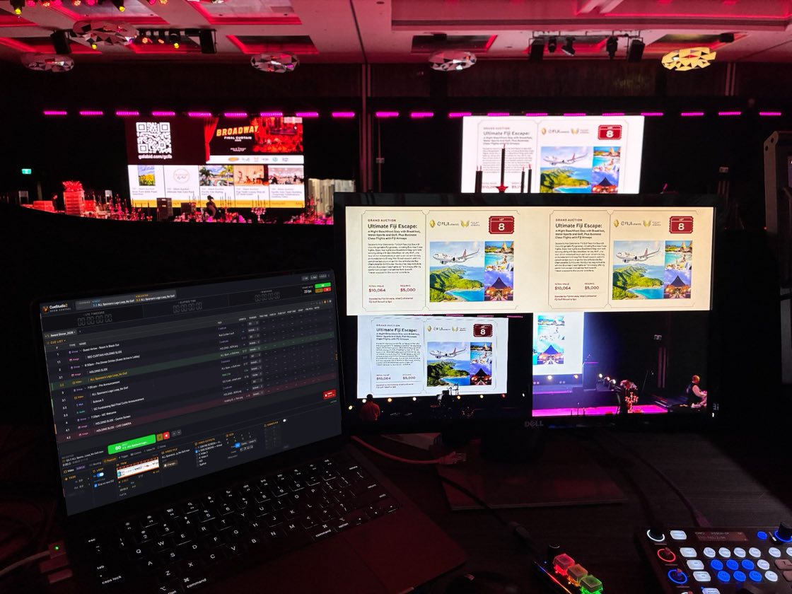 A darkened function room during a gala dinner. A MacBook on the tech table runs a CueStudio cue list; the monitor beside it shows the auction lot graphic being sent to the room's screens next to a live camera feed of the stage, with the room's screens and magenta house lighting behind.